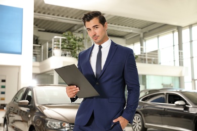 Young salesman with clipboard near car in modern dealership