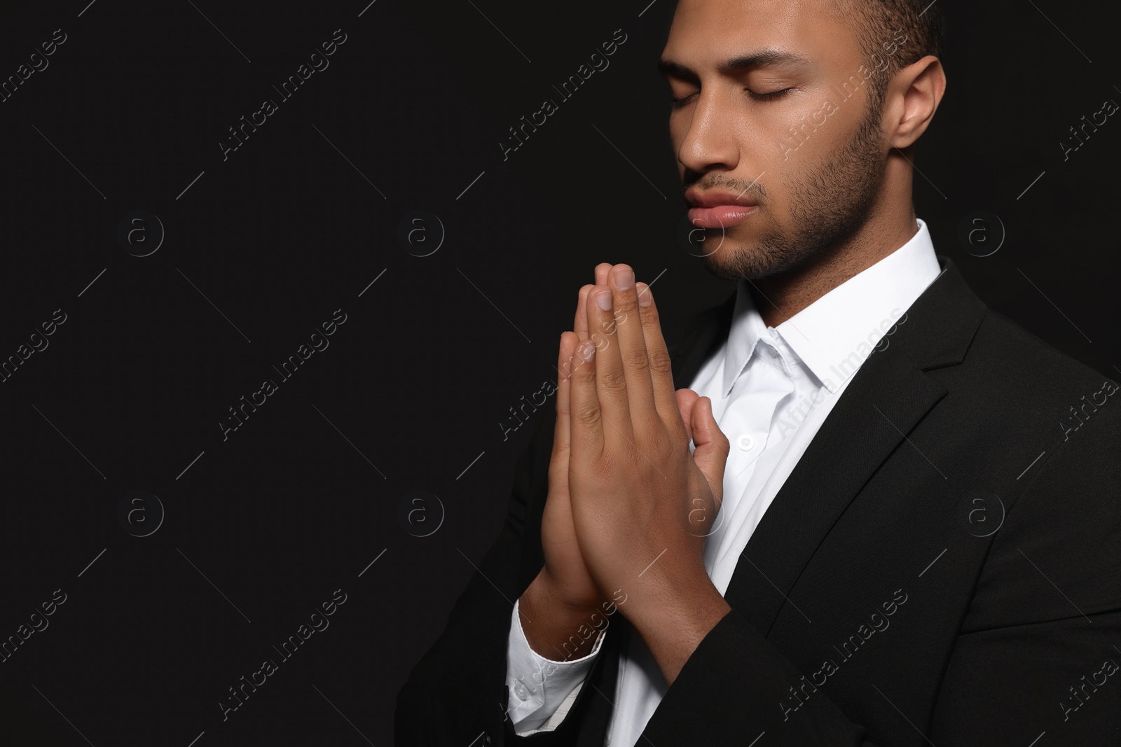 Photo of African American man with clasped hands praying to God on black background. Space for text