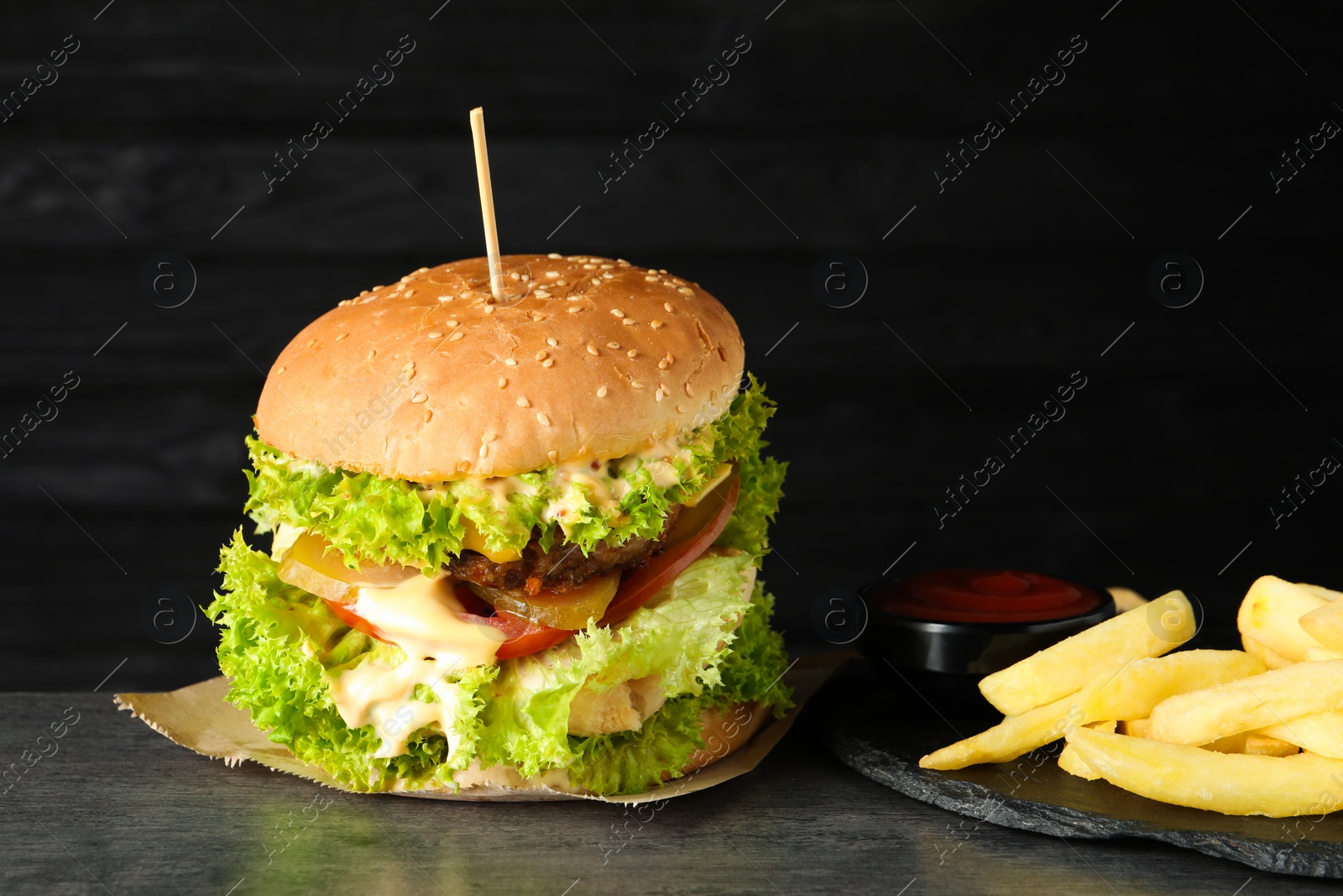 Burger with delicious patty, french fries and sauce on gray table against dark background Photo of Burger with delicious patty, french fries and sauce on gray table against dark background