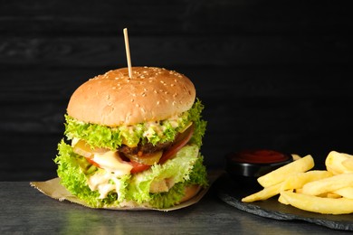 Photo of Burger with delicious patty, french fries and sauce on gray table against dark background