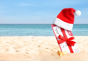 Image of Ticket with red bow and Santa hat on sandy beach, space for text. Christmas vacation