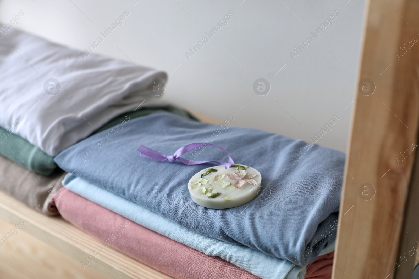 Photo of Stacked clothes and beautiful scented wax sachet on shelf near white wall, closeup