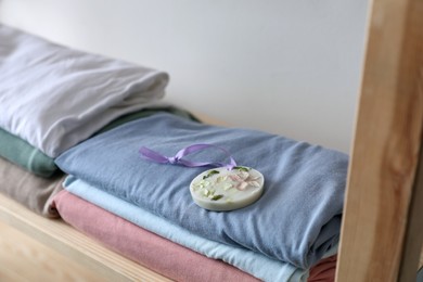 Photo of Stacked clothes and beautiful scented wax sachet on shelf near white wall, closeup