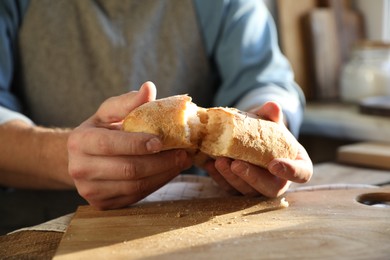 Photo of Man breaking loaf of fresh bread at wooden table indoors, closeup