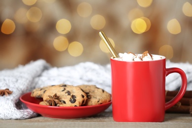 Cup of tasty cocoa with marshmallows and cookies on wooden table against blurred lights