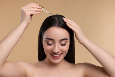 Happy young woman applying essential oil onto hair roots on beige background, closeup