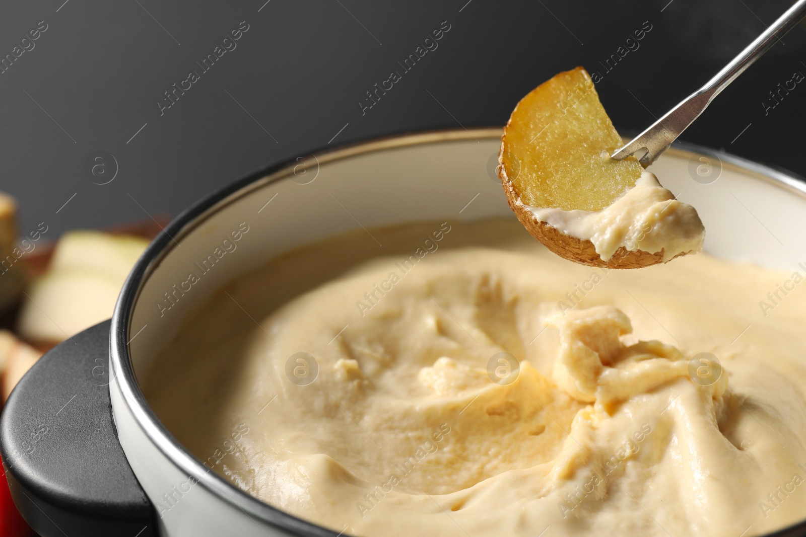 Photo of Dipping piece of potato into fondue pot with melted cheese on grey background, closeup