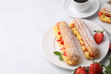 Photo of Delicious eclairs filled with cream, strawberries and mint on white tiled table, space for text