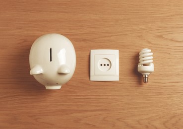 Photo of Piggy bank, power socket and fluorescent light bulb on wooden table, flat lay. Energy saving concept