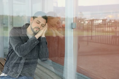 Tired man sleeping at public transport stop outdoors, view through glass. Space for text