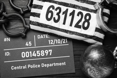 Photo of Metal ball with chain, prison uniform and mugshot letter board on black wooden table, flat lay