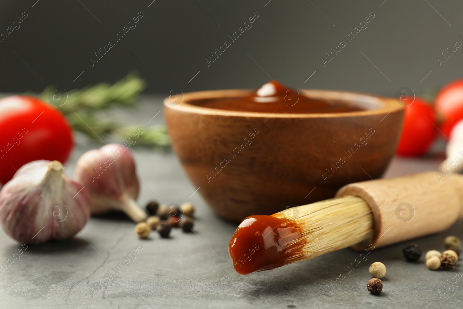 Photo of Brush with tasty barbeque sauce, bowl, garlic and peppercorns on grey table, closeup