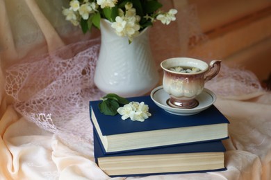 Photo of Cup of aromatic tea, beautiful jasmine flowers and books on fabric indoors