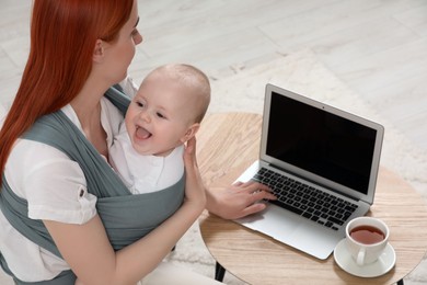 Mother using laptop while holding her child in sling (baby carrier) indoors