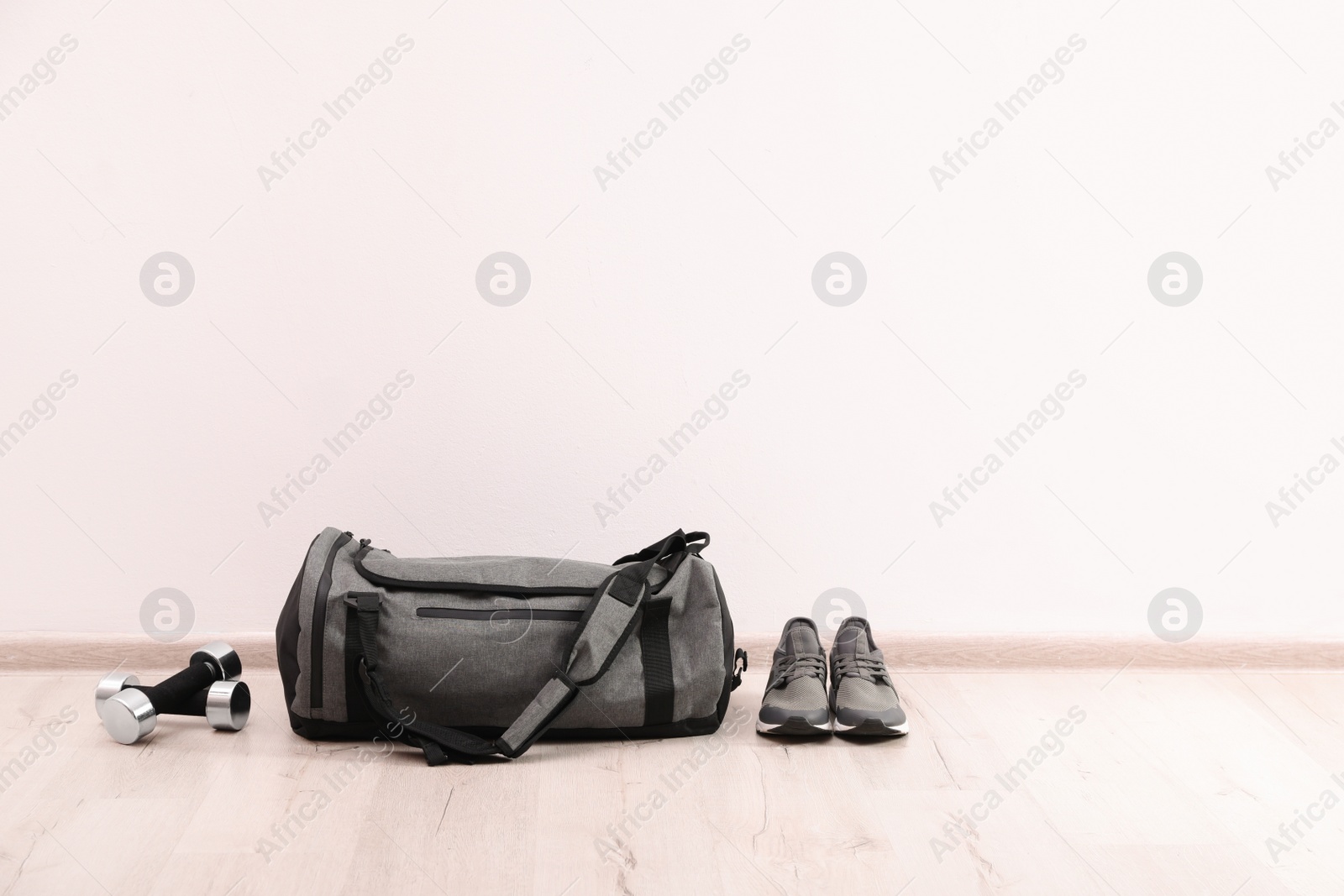 Photo of Grey sports bag, sneakers and dumbbells on floor near white wall, space for text