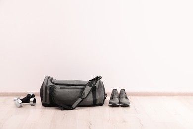 Grey sports bag, sneakers and dumbbells on floor near white wall, space for text