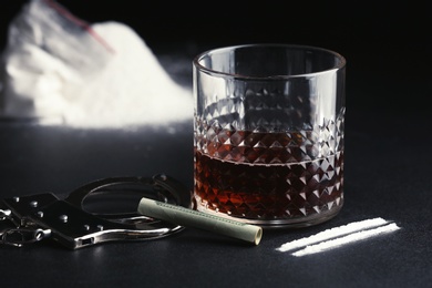 Photo of Glass of alcohol, cocaine, money and handcuffs on dark table