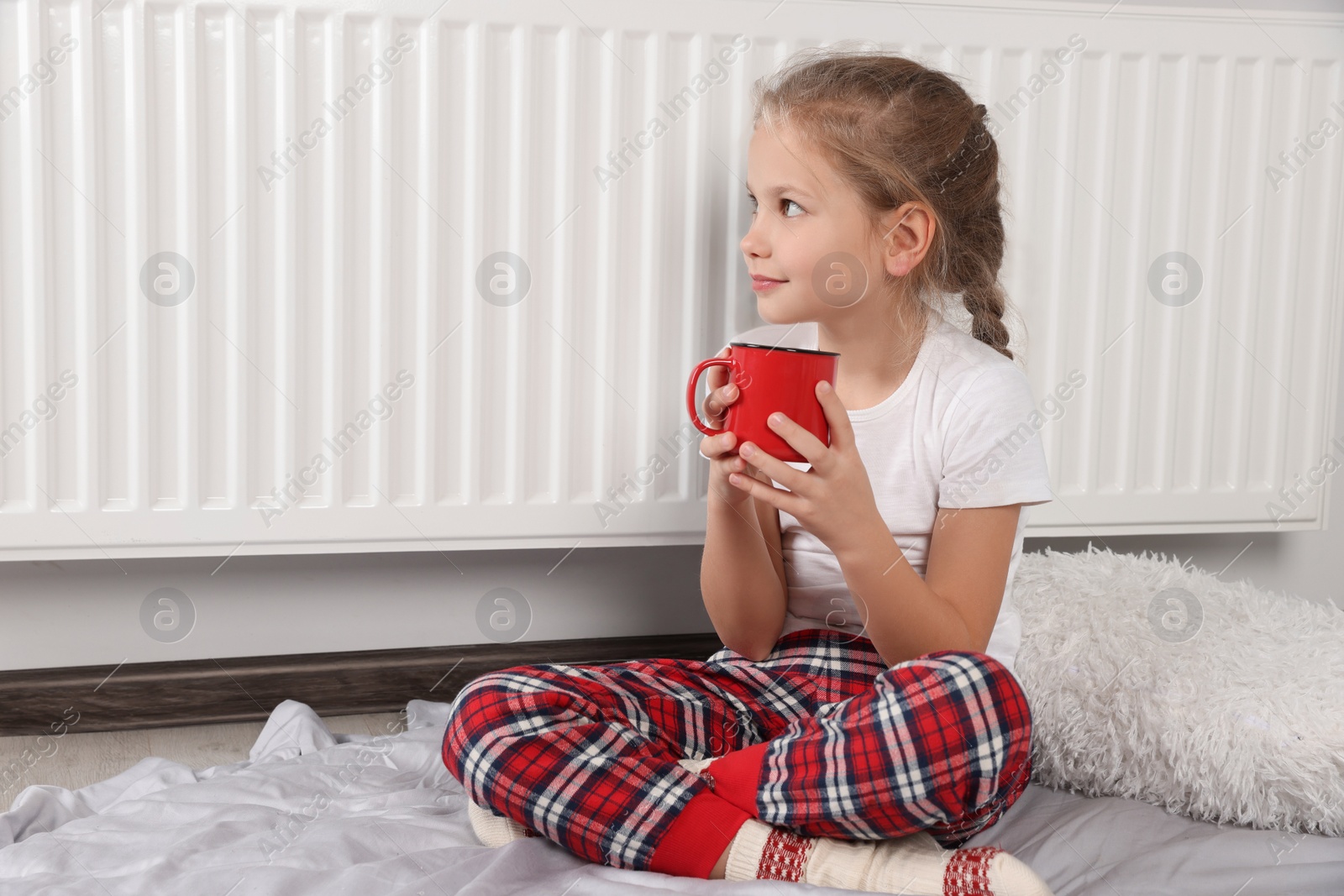 Photo of Little girl with cup of hot drink near heating radiator indoors, space for text