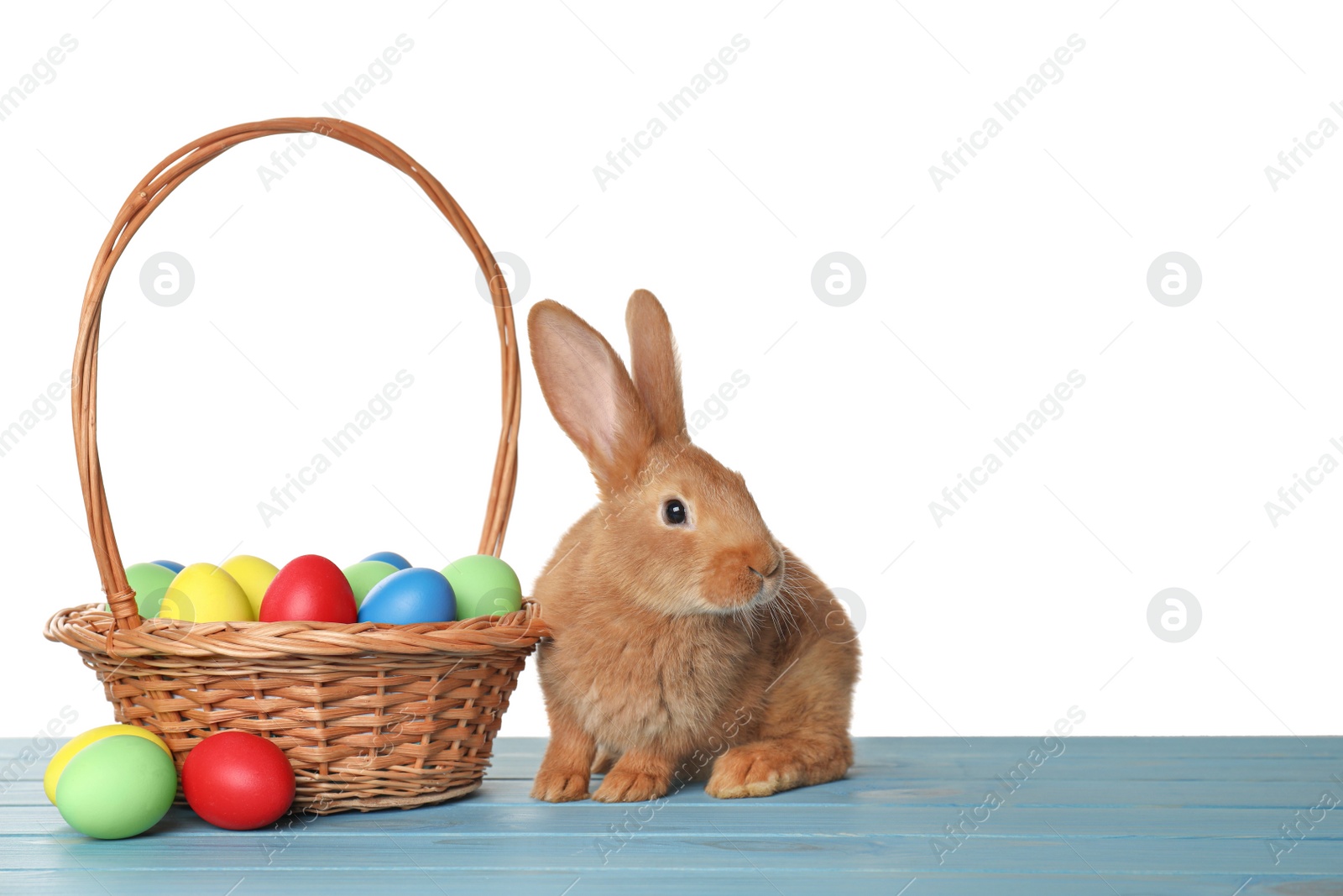 Photo of Cute fluffy bunny and wicker basket with Easter eggs on light blue wooden table