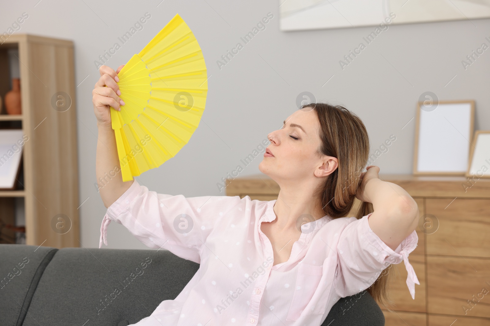 Photo of Woman waving yellow hand fan to cool herself on sofa at home