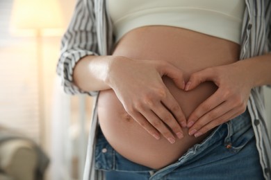 Photo of Pregnant woman making heart with her hands near belly indoors, closeup