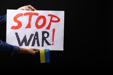 Photo of Woman holding Ukrainian flag and poster with words Stop War on black background, closeup