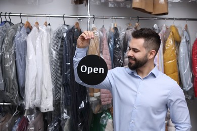 Photo of Dry-cleaning service. Happy worker holding Open sign near clothes in plastic bags indoors