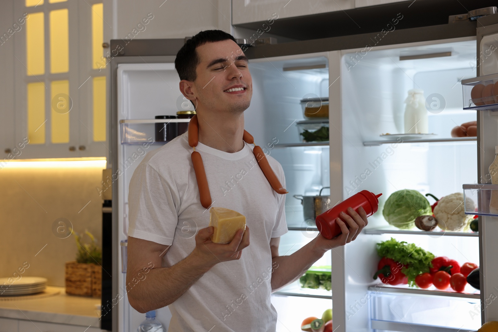 Photo of Happy man with ketchup, cheese and sausages near refrigerator in kitchen