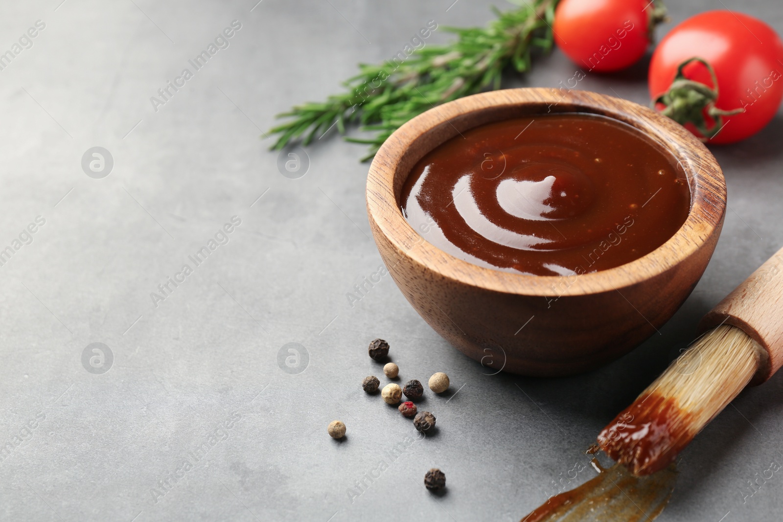 Photo of Tasty barbeque sauce in bowl, brush and peppercorns on grey textured table, closeup. Space for text