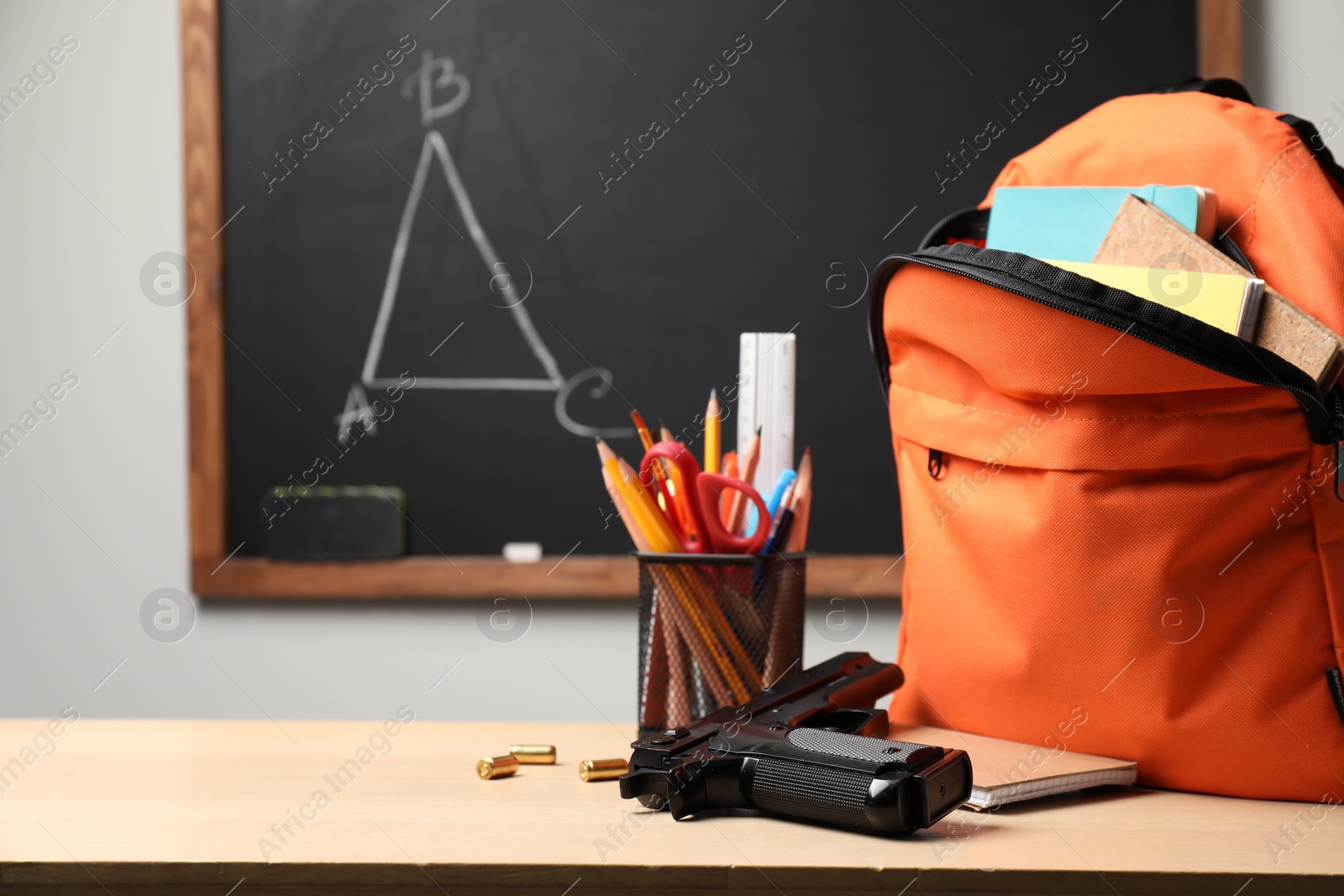 Photo of Gun, bullets and school stationery on wooden table near blackboard indoors, space for text