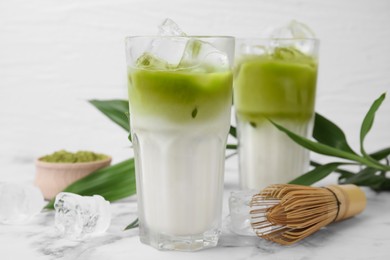 Glasses of tasty iced matcha latte, bamboo whisk and leaves on white marble table, closeup