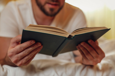 Photo of Young man reading book on bed at home, closeup