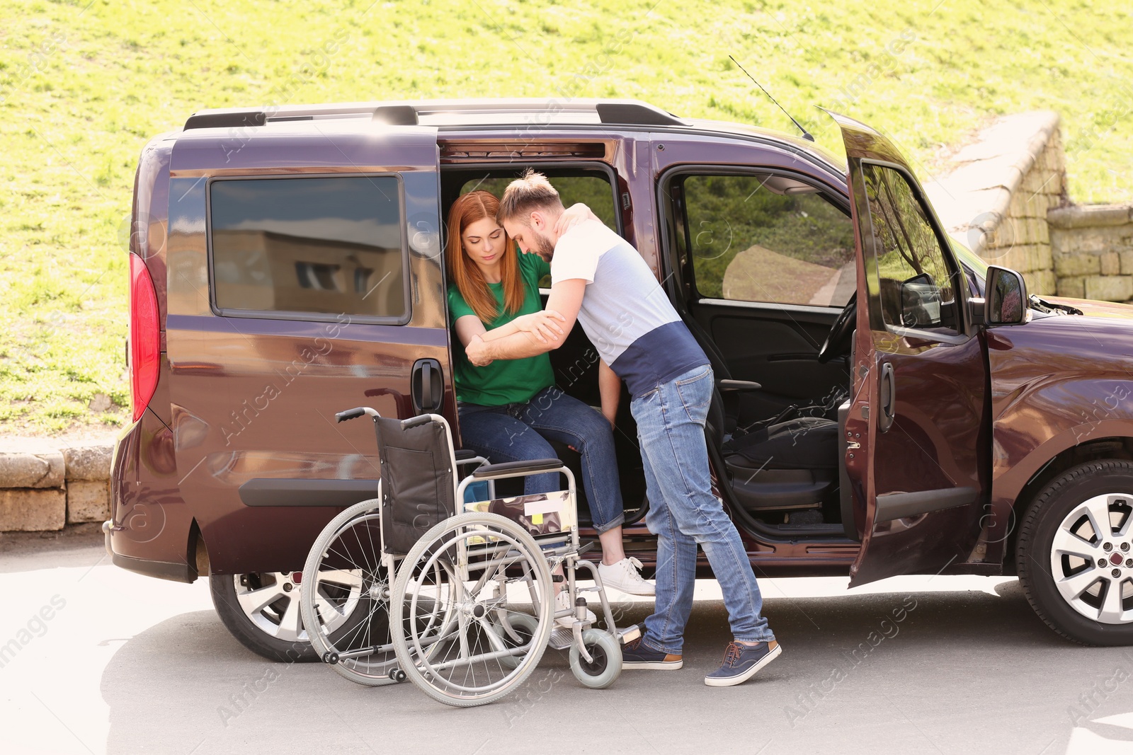 Photo of Young man helping woman to get out from van into wheelchair outdoors