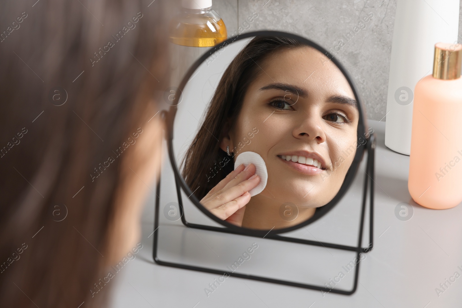 Photo of Young woman cleaning her face with cotton pad near mirror at light grey table indoors