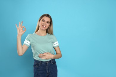 Photo of Happy woman touching her belly and showing okay gesture on light blue background, space for text. Concept of healthy stomach