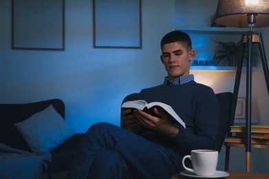Photo of Young man with cup of drink reading book in cozy room at night