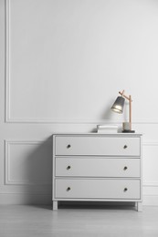 Photo of Lamp and books on chest of drawers near white wall indoors