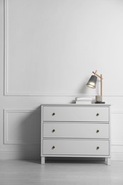 Lamp and books on chest of drawers near white wall indoors