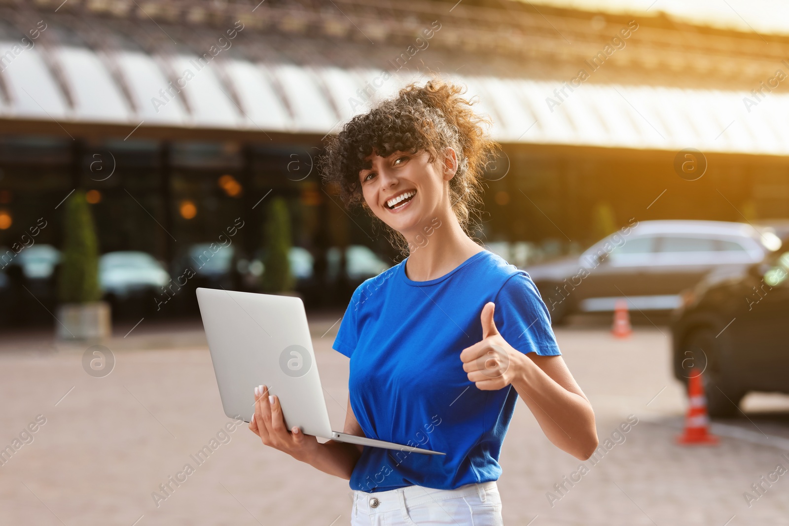 Photo of Happy young woman using modern laptop and showing thumb up on city street