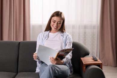 Photo of Happy woman reading magazine on sofa with wooden armrest table at home