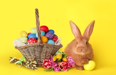 Image of Cute bunny and wicker basket with bright Easter eggs on yellow background