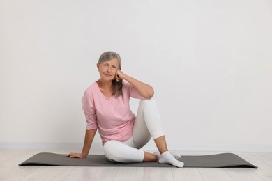 Senior woman sitting on mat near white wall, space for text. Yoga practice