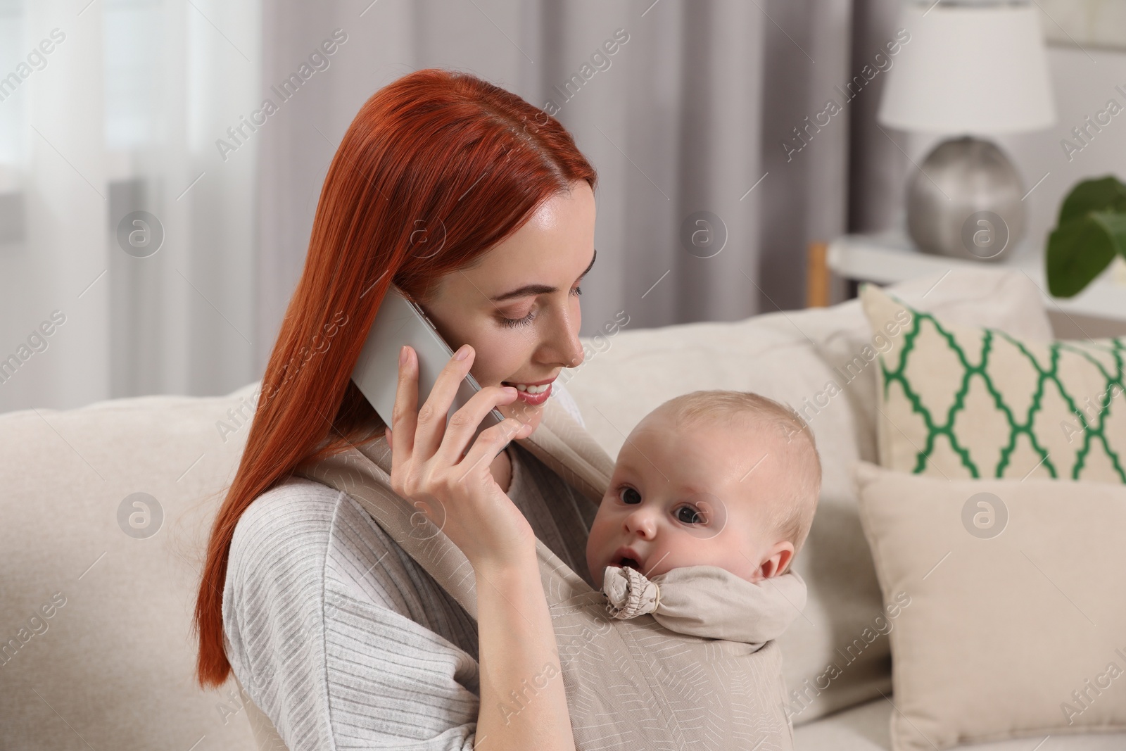 Photo of Mother talking on smartphone while holding her child in sling (baby carrier) at home