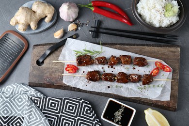 Skewers with pieces of tasty chicken meat glazed in soy sauce and other products on grey table, flat lay