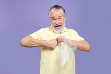 Photo of Emotional senior man popping bubble wrap on light purple background. Stress relief