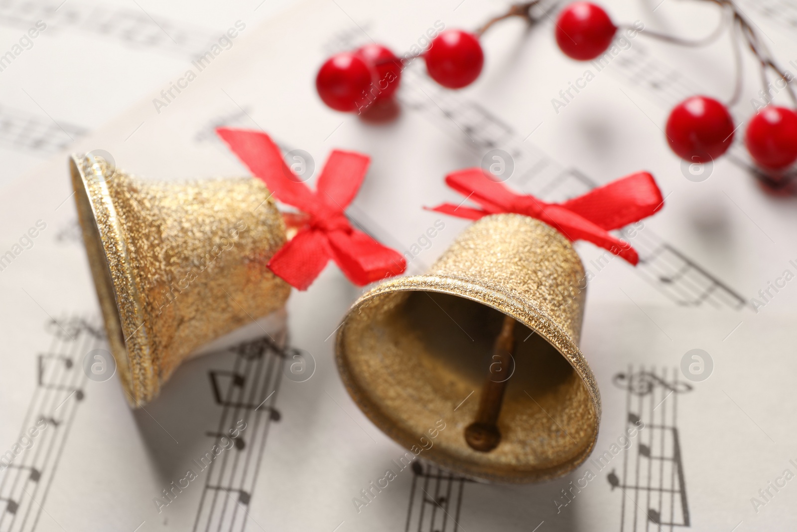Photo of Golden shiny bells with red bows and decorative berries on music sheets, closeup. Christmas decoration
