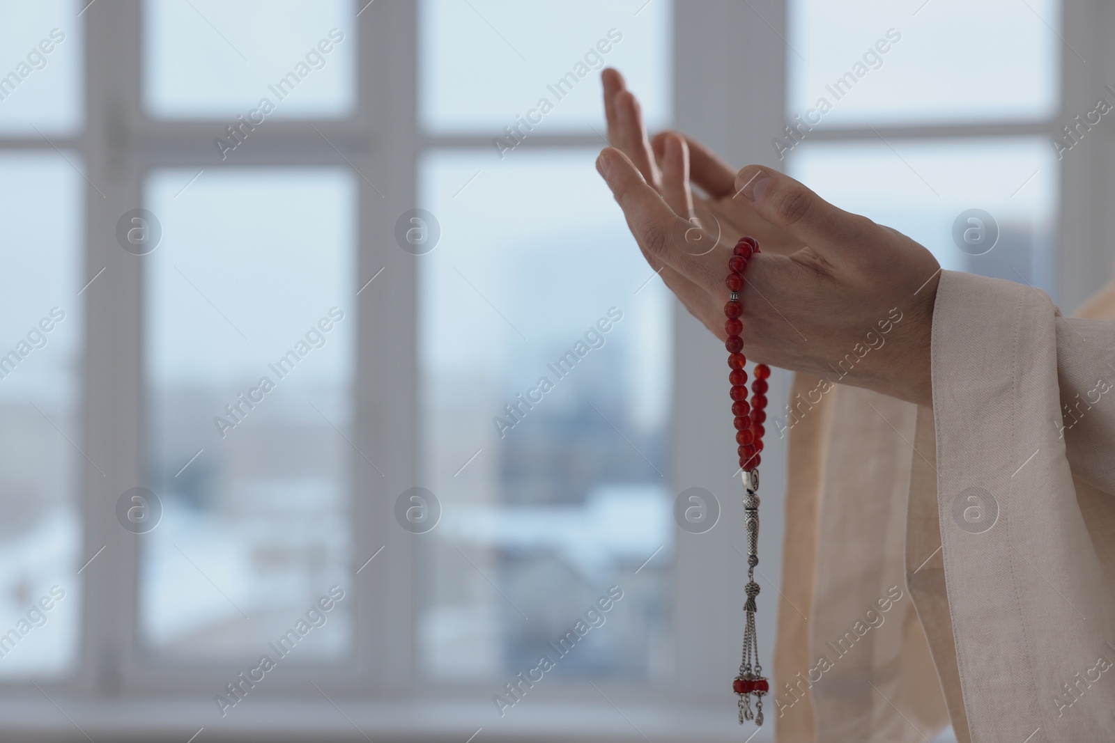 Photo of Muslim man with misbaha praying near window indoors, closeup. Space for text