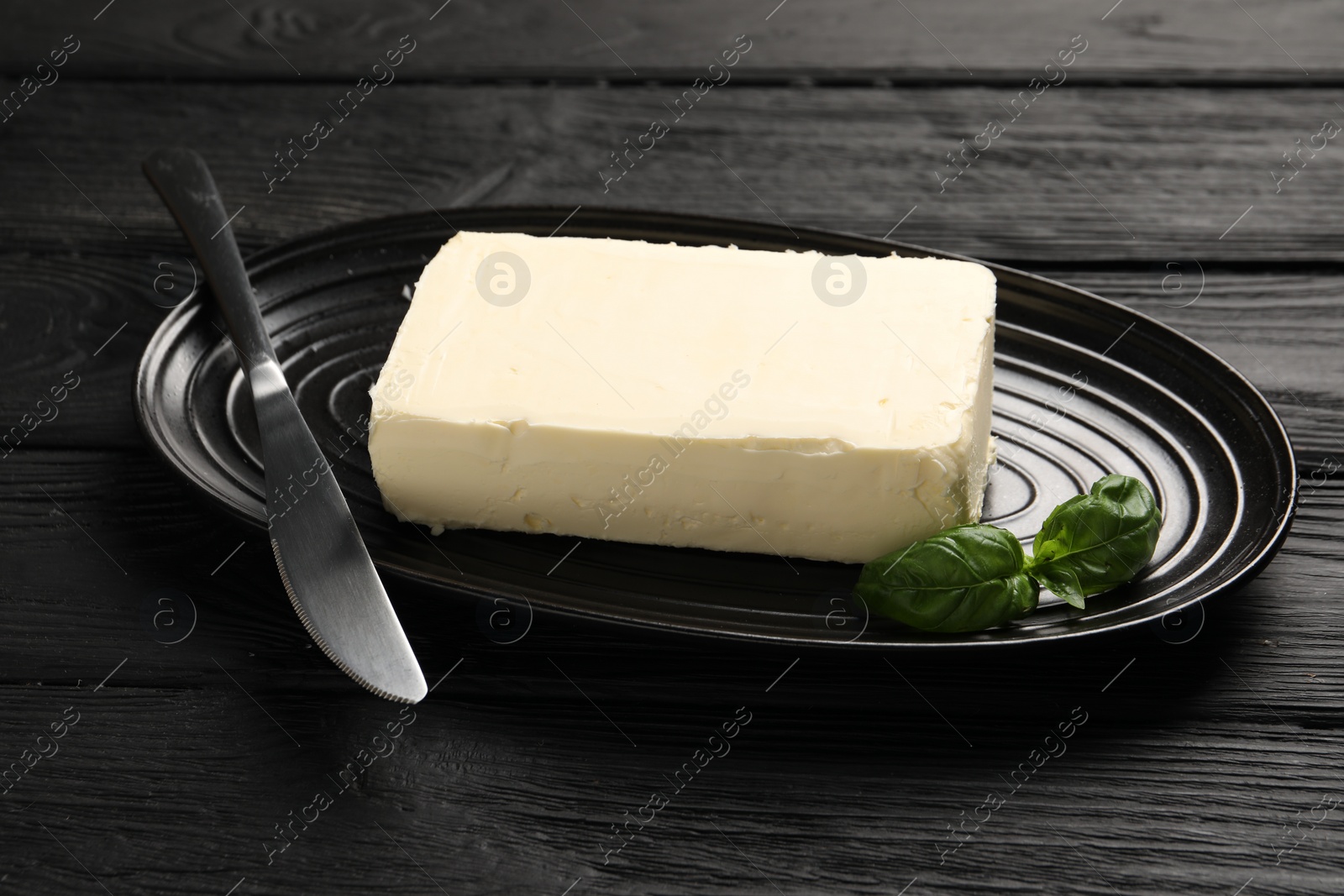 Photo of Block of tasty butter with basil and knife on black wooden table