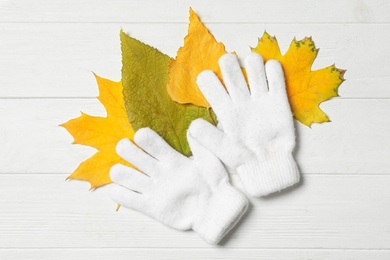 Stylish woolen gloves and dry leaves on white wooden table, flat lay