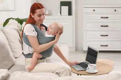 Photo of Mother holding her child in sling (baby carrier) while using laptop at home